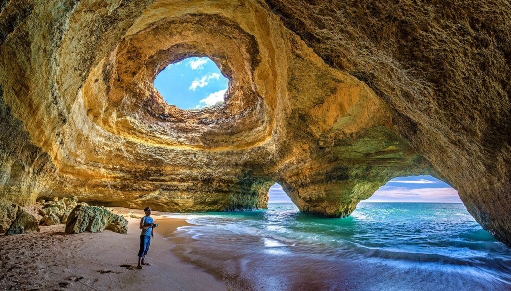 This image features a beautiful cave on the Algarve. A man stands with his camera with crystal waters around him and an opening in the cave showing the blue sky and clouds.