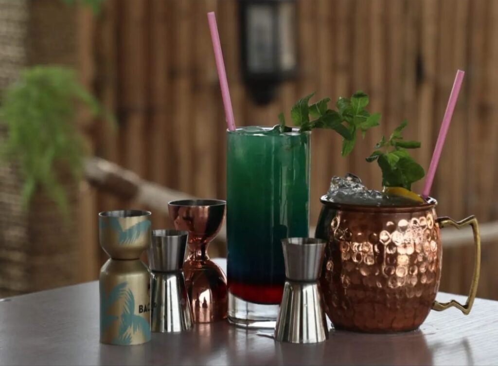 A refreshing cocktail mix in a glass, garnished with fresh mint leaves hanging from the rim, accompanied by a mug of ice cubes and a selection of shot mixers.