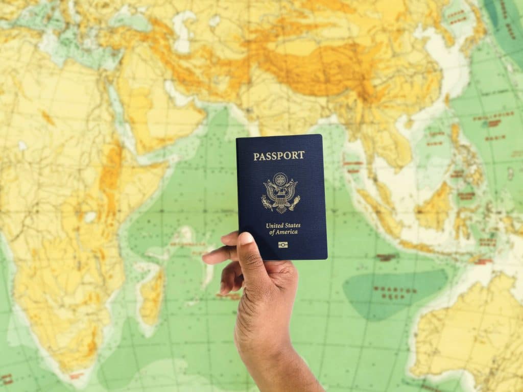 A hand holding an American passport is raised in front of a world map, symbolizing the privilege of passport and the freedom of travel it represents, highlighting the global access and opportunities afforded to its holder.