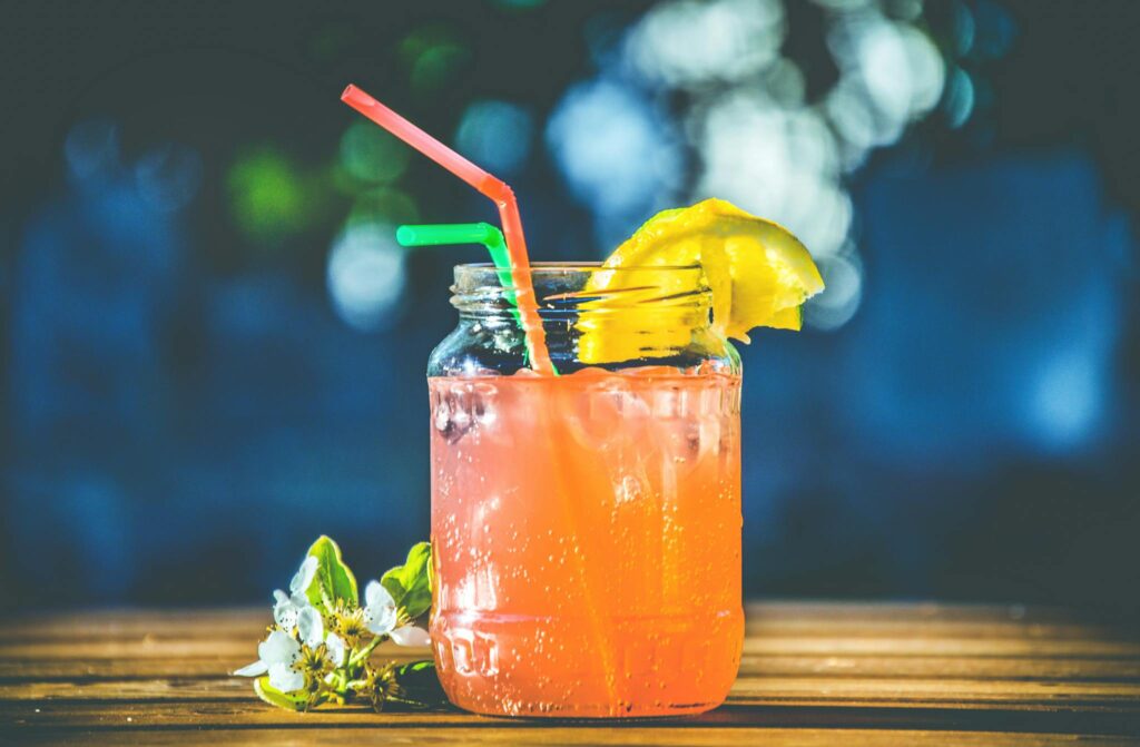 A refreshing drink composed of an alcoholic beverage poured into a clear glass, with two straws placed alongside each other inside the glass. A slice of juicy orange is perched on the rim of the glass, adding a touch of colour to the drink.